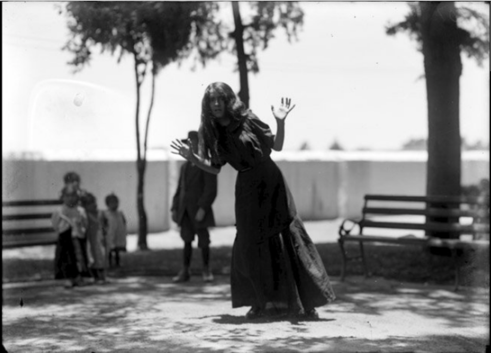 A grainy black and white photo of a woman in a long dress swaying at the waist with her hands up by her face