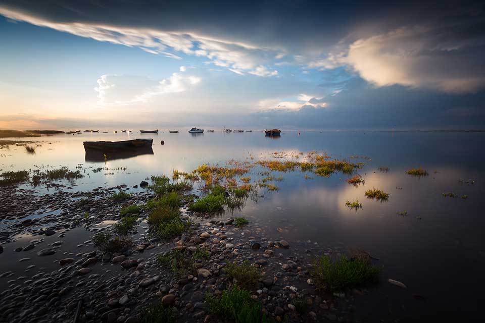 A boat floats in a desultory natural port as the sun begins to crest above the horizon