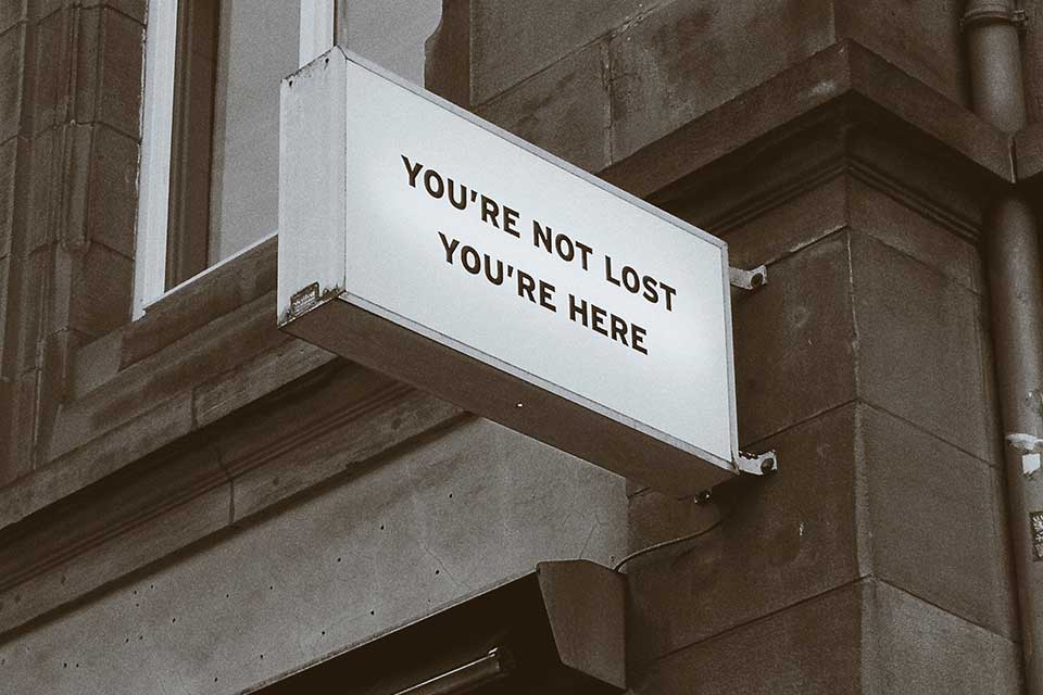 A sign mounted on the corner of a building. The sign reads, ”You are not lost. You are here.”