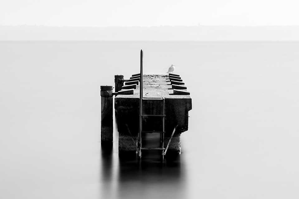 A black and white photo of a small boat dock surrounded by water, which stretches unbroken to the horizon