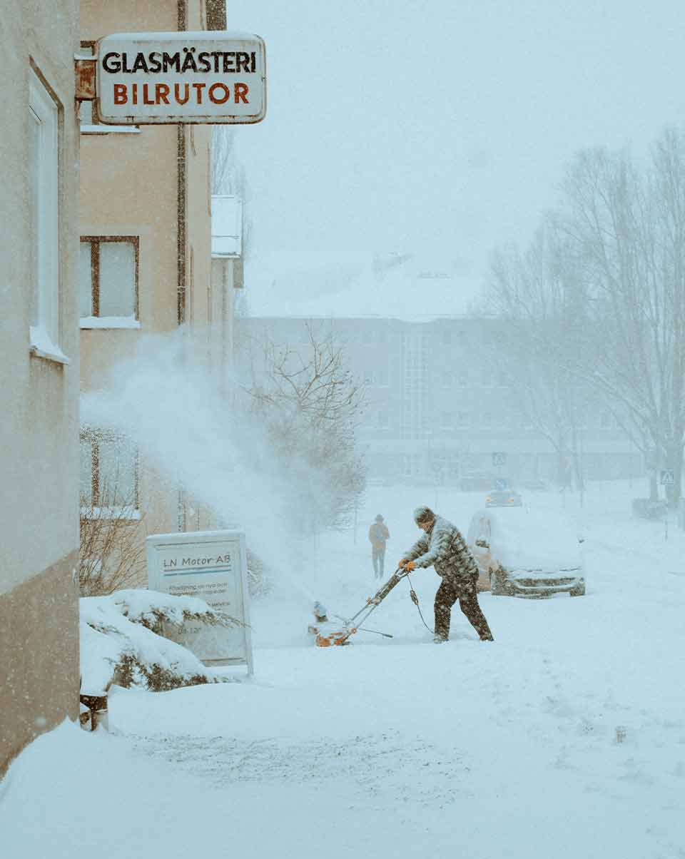 A photograph of a man running a snow blower on a city street