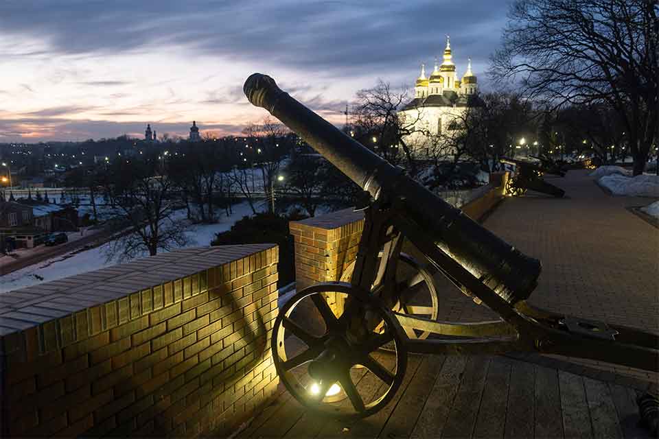 A photograph of a cannon perched atop a roof at night, overlooking a subdued snowy city landscape