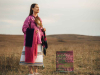 A photograph of a woman in indigenous garb standing on scrub grass hilltop with a painting nearby