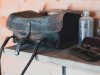 A weathered leather bag sits on a dresser with a dusty glass bottle and a pair of antique glasses sitting nearby