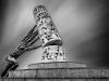 A black and white photo of a human form with many arms but no head pushing a column adorned in cuneiform back on to its pedestal.