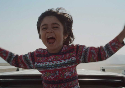 A still image of a boy with his arms outstretched coming out of the top of a moving car